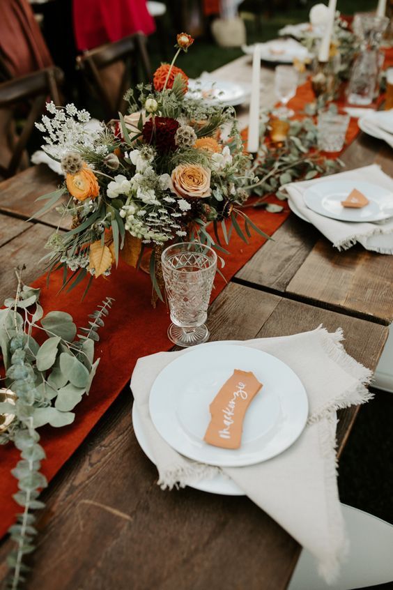 a boho fall wedding tablescape with a rust suede table runner, white napkins, bold yellow and rust blooms and greenery