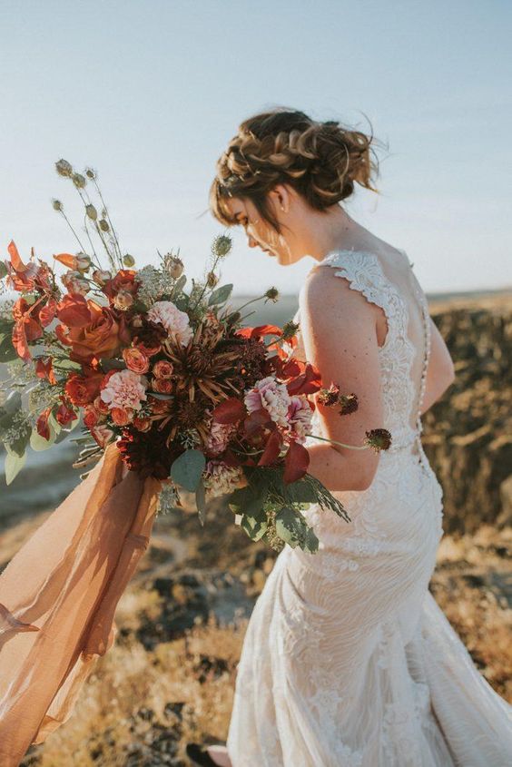 a bold and lush fall wedding bouquet of pink, rust, deep red and deep purple blooms, greenery, seed pods and long ribbons is amazing