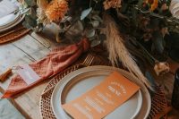 a bold retro inspired wedding tablescape with a woven placemat, neutral plates, orange napkins, a bold orange and neutral bloom centerpiece with greenery