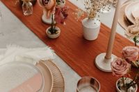a bold wedding tablescape with a rust-colored table runner, neutral candles, pink blooms, blush menus and wooden cutlery