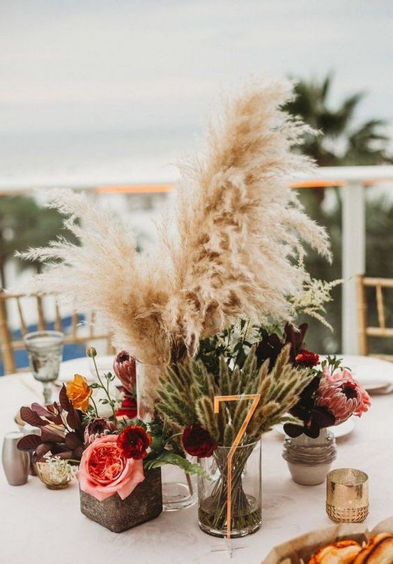 a bright fall centerpiece of bold blooms, greenery, pampas grass and a gold table number in front of wheat