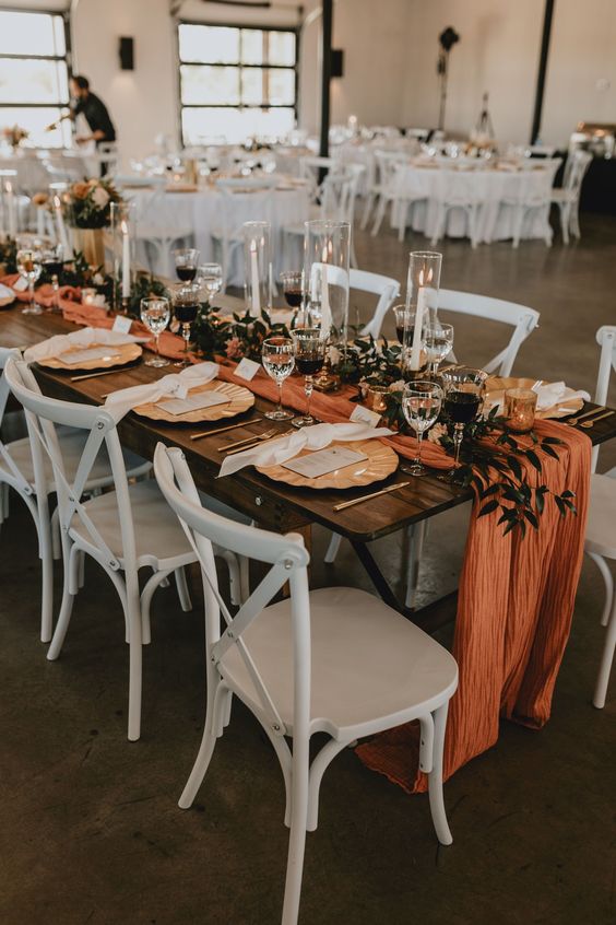 a bright fall wedding tablescape with an orange runner, greenery and candlees, gold chargers and neutral napkins is a lovely idea for the fall