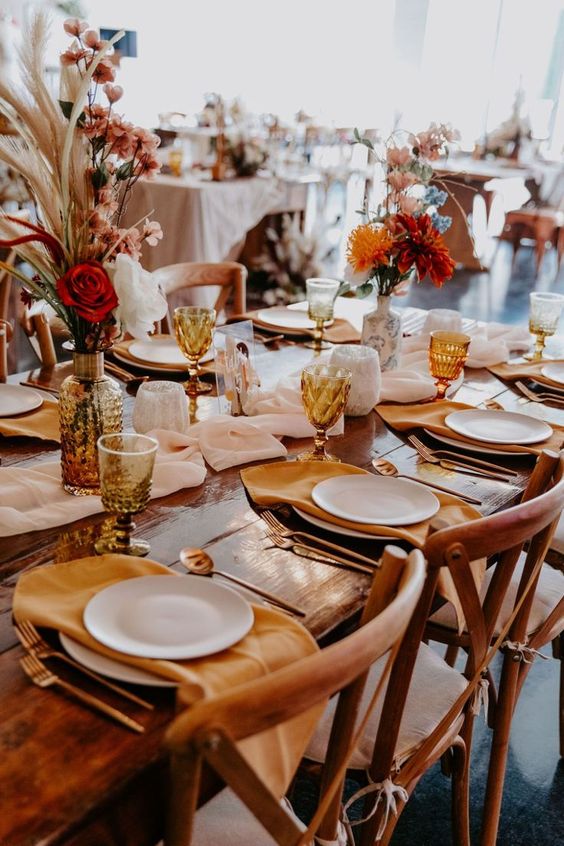 a bright modern wedding tablescape with amber napkins, glasses and vases, red and rust blooms and grasses