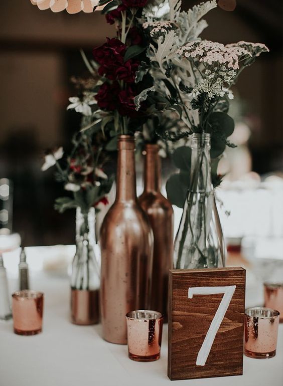 a chic and simple wedding centerpiece of copper candleholders and bottles as vases for greenery and dark and white blooms