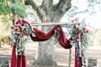 a chic fall wedding arch of burgundy velvet, greenery and burgundy blooms is very refined