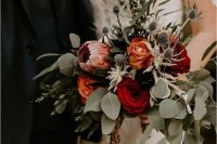 a colorful fall wedding bouquet of orange, deep red and purple blooms, a pink king protea, greenery and thistles is gorgeous