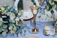 a copper placemat and cutlery, a gold candlehoder and white and blue blooms and greenery