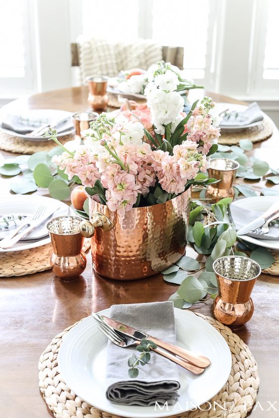 a cozy tablescape with copper hammered cups, a vase and peachy pink blooms plus wicker chargers