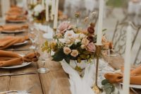 a delicate fall wedding tablescape with a neutral runner, rust napkins, pretty floral arrangements, greenery and candles in gold candleholders