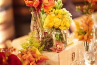 a fall vineyard table decoration of a crate, bright blooms in vases, moss and a table number with nails