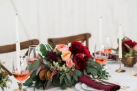 a fall wedding centerpiece of lush greeneyr, blush and peachy blooms, burgundy flowers and white candles