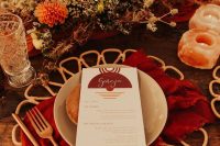 a fantastic boho wedding tablescape with a bold orange, rust, burgundy, peachy floral centerpiece, quartz candleholders, red napkins and rattan placemats