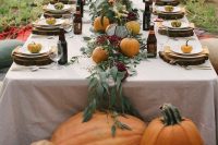 a leafy table runner with burgundy blooms and fresh pumpkins, wood slices, plates, pumpkins and beer bottles for a fall wedding