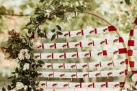 a lovely flal or winter seating chart of an oversized embroidery hoop, greenery and neutral blooms, cards with berries attached to red fabric straps