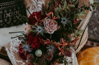 a lush and moody fall wedding bouquet with burgundy, blush, rust blooms, lots of greenery and blue thistles, with much dimension and texture featured