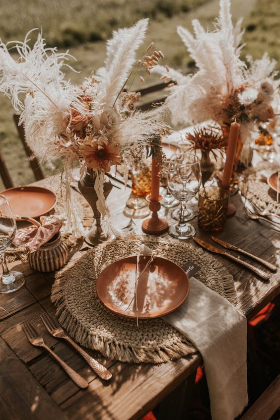 a lush boho wedding tablescape with copper plates, candles and blooms, cutlery, raffia placemats, neutral linens and dired grasses