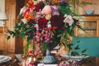 a lush fall wedding centerpiece of a vintage urn, red, burgundy, purple, orange and pink blooms, greenery and grapes