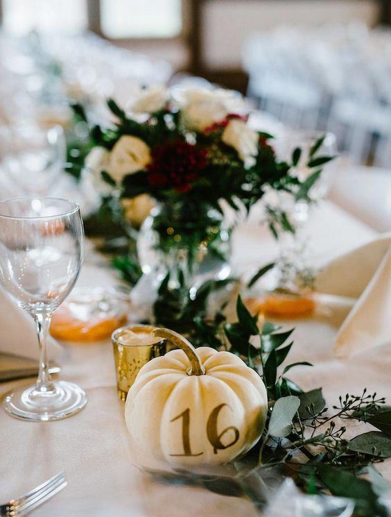 a mini pumpkin with a table number, greenery and candles for fall wedidng table styling