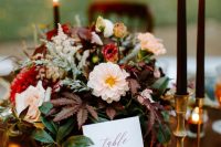 a moody fall wedding centerpiece of red and blush blooms, foliage and herbs and black candles