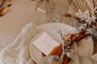 a neutral fall wedding table setting with white plates, jute placemats, neutral and orange napkins, dried blooms, leaves and fronds and candles