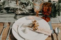 a pretty and catchy fall wedding tablescape with an olive green table runner, neutral porcelain, elegant cutlery, greenery, amber glasses and tall candles
