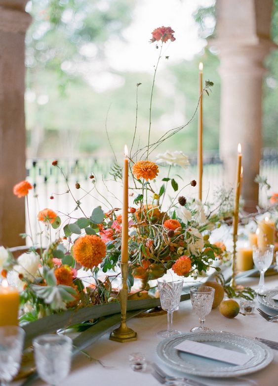 a pretty fall wedding centerpiece of white and orange blooms, greenery and orange candles is refined and chic