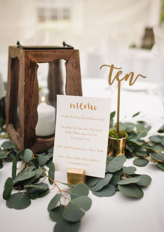 a simple wedding centerpiece of greenery, a wooden candle lantern, a calligraphy table number in a shot with moss