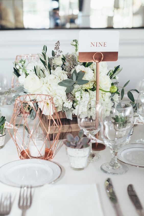 a stylish rustic centerpiece of a wooden crate with white blooms and greenery, dark succulents, a copper candleholder and a copper and white table number