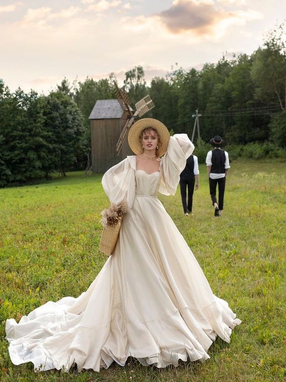 a vintage-inspired A-line ivory wedding dress with a corset, puff sleeves, a train, a pearl choket and earrings, a straw hat and a bag