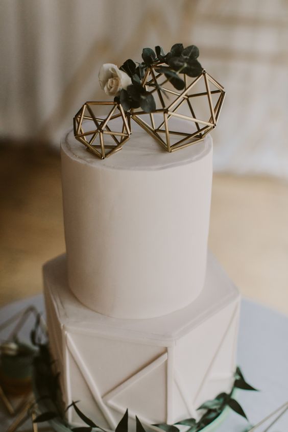 a white geometric wedding cake with gold himmeli toppers and greenery and white blooms