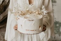 a white semi-naked wedding cake topped with white dried blooms and leaves for a boho wedding