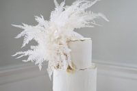 a white textural wedding cake with a gold ege and lush white dried leaves is a cool wedding dessert
