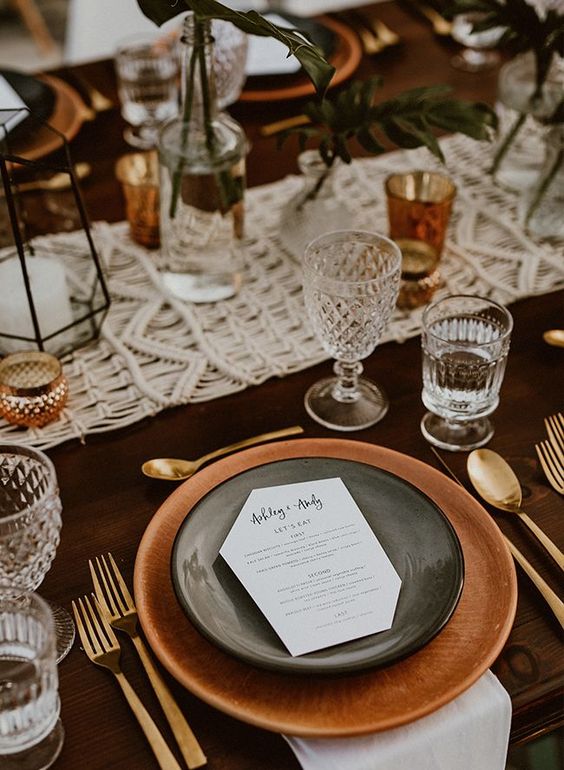 an elegant boho wedding tablescape with terracotta plates, amber glasses and candleholders, a macrame runner and candle lanterns