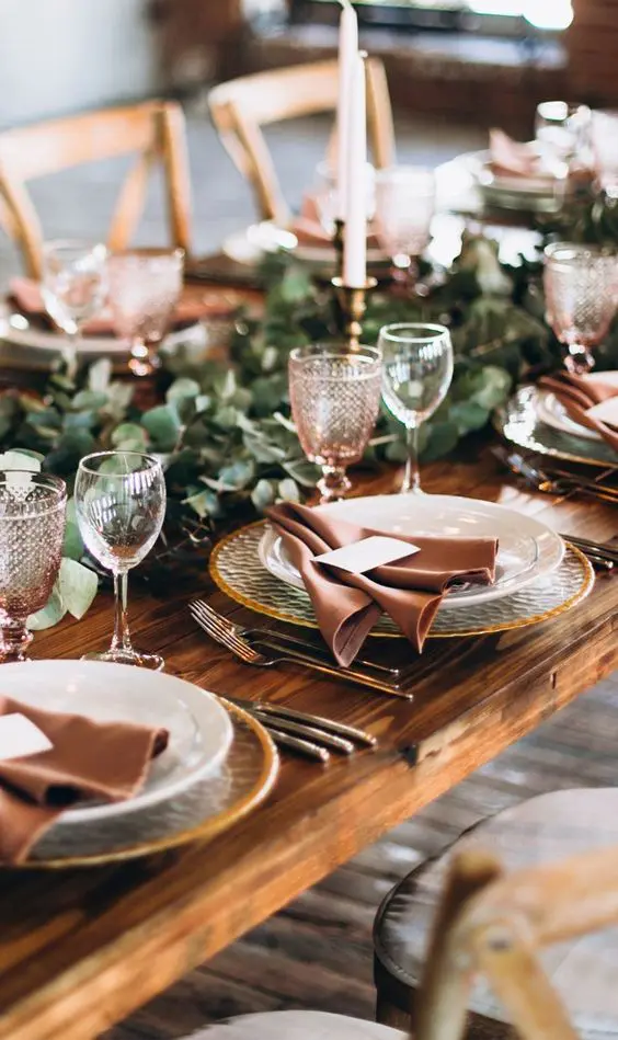 an elegant rustic fall tablescape with a greenery runner, tall candles in gold candlesticks, hammered chargers, pink glasses and dusty pink napkins