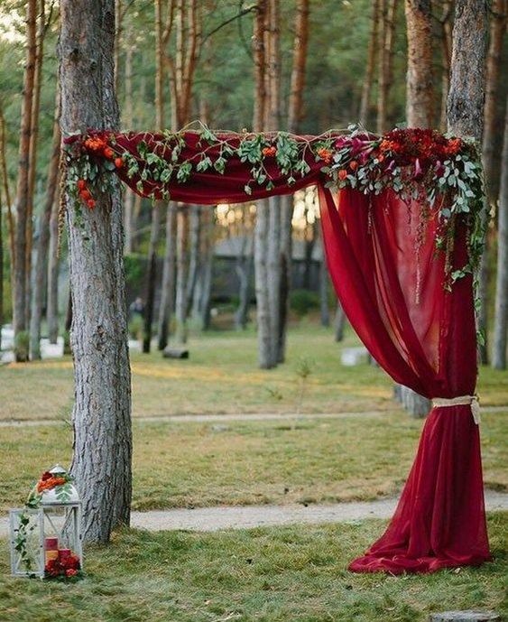 an outdoor fall wedding altar with a red curtain, greenery and bold blooms plus a candle lantern with red candles