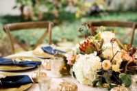 an outdoor fall wedding table with a lush floral centerpiece, gold chargers, navy napkins with gold cutlery and gold pumpkins for decor