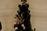 a black wedding cake with black skulls, burgundy and white blooms and gilded leaves plus a gold skull on top