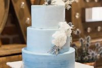 a blue watercolor ombre wedding cake topped with white blooms and thistles on top is a chic and dreamy idea for a wedding