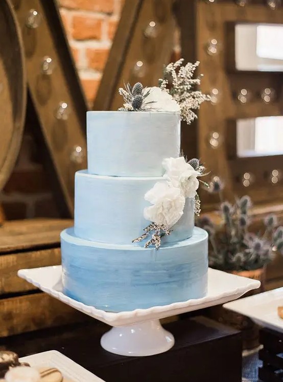 a blue watercolor ombre wedding cake topped with white blooms and thistles on top is a chic and dreamy idea for a wedding