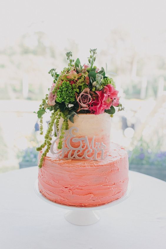 a cheerful ombre pink textural buttercream wedding cake with lots of bright blooms and greenery on top and a calligraphy topper is wow