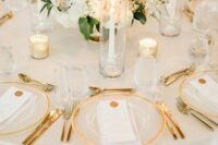 a chic wedding tablescape in white and gold, with a white floral centerpiece and tall and thin candles, clear chargers and gold cutlery
