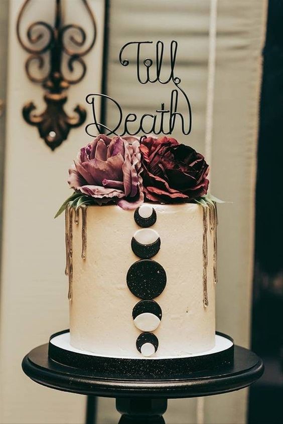 a cool Halloween wedding cake with moon phases, pink and burgundy blooms, gold drip and a calligraphy topper
