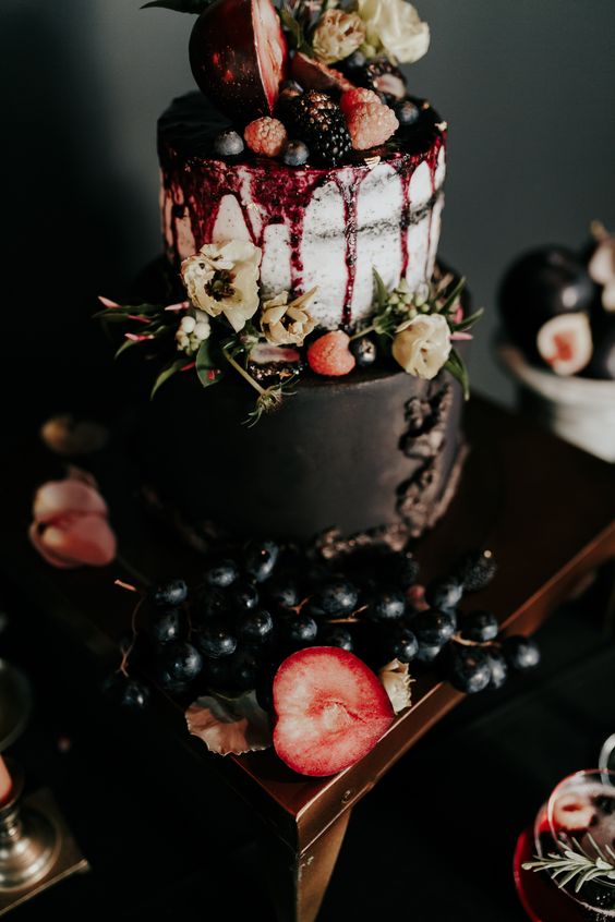 a decadent wedding cake with a black and a naked tier, berry drip, berries and fruits plus blooms on top