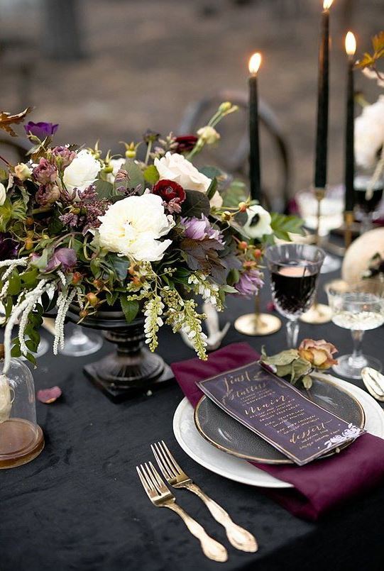 a jewel-tone Halloween wedding tablescape with a black tablecloth, a purple napkin, purple and white blooms and greenery and black candles