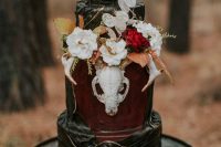 a moody Halloween wedding cake in black and burgundy, with white and bright blooms, dried leaves, patterns and a sugar skull