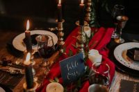 a moody luxurious wedding tablescape with a red table runner, black candles, gold touches and greenery