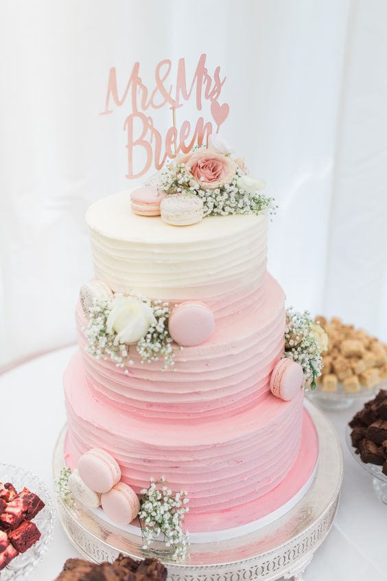 a pretty rustic wedding cake in ombre pink, with textural buttercream, pink and neutral macarons, white and pink blooms and a calligraphy topper