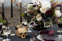 a refined Halloween wedding table with a black tablecloth and candles, burgundy napkins, a chic floral centerpiece, gold cutlery and a skull