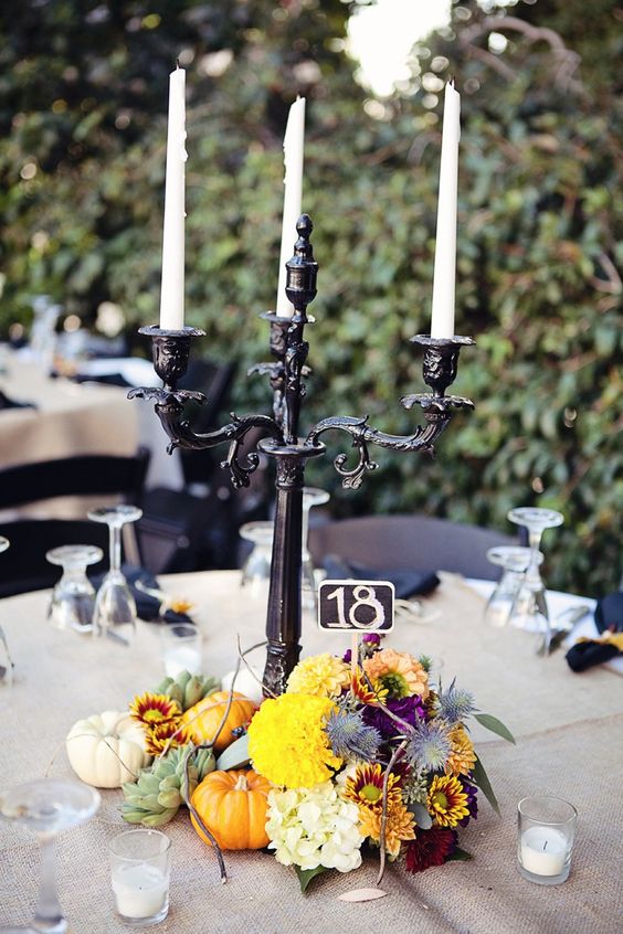 a rustic Halloween wedding tablescape with a burlap tablecloth, a black candelabra, bright blooms, pumpkins, thistles and candles around