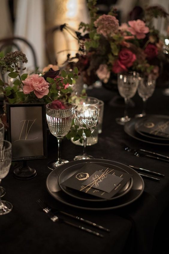 a sophisticated Halloween wedding tablescape with a black tablecloth, plates and cutlery, moody florals and greenery plus candles